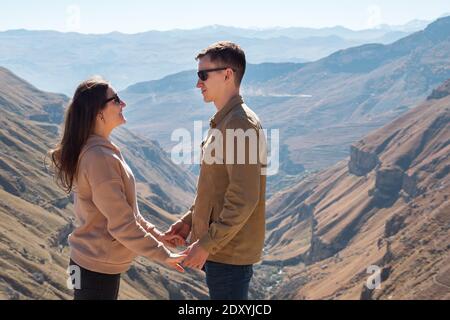 La brunetta con lunghi capelli sciolti tiene le mani dell'uomo dentro occhiali da sole in piedi contro profondo canyon tra alte montagne sotto luminoso luce solare Foto Stock