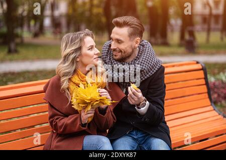 L'uomo abbracciante ha trovato una piccola coppia felice di foglia seduta sulla panchina abbracciata in parco indossando cappotti e sciarpe raccogliendo un bouquet di foglie cadute. Amore Foto Stock