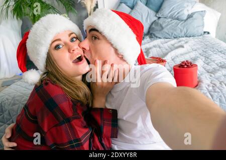 Festa di Natale online. Uomo mordente donna prendendo selfie indossando cappelli di santa e pigiami rossi sullo sfondo dei regali di Capodanno Foto Stock