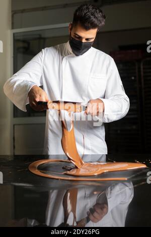 Giovane pasticciere con maschera che lavora sul cioccolato temperante. Foto Stock