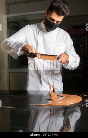 Giovane pasticciere con maschera che lavora sul cioccolato temperante. Foto Stock