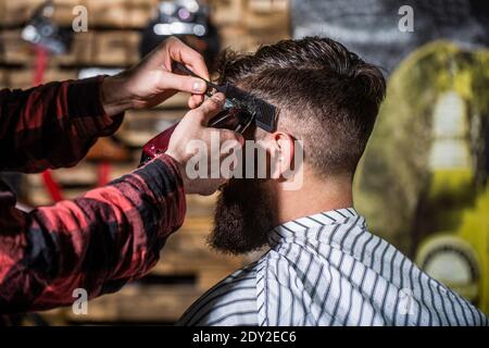 Barbiere funziona con regolacapelli. Cliente hipster ottenere haircut. Mani di barbiere con regolacapelli, primo piano. Uomo bearded in barbiere. Taglio di capelli Foto Stock