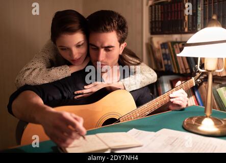 Giovane donna abbracciando il ragazzo durante la lettura delle note in blocco note processo di creazione di canzoni nel vecchio appartamento in serata Foto Stock