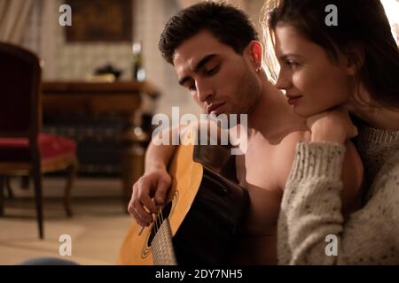 Vista laterale del giovane hugging femminile ha messo a fuoco il gioco del giovane uomo chitarra durante la data romantica in serata tranquilla a casa Foto Stock