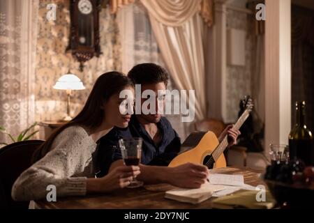 Giovane con chitarra e donna con note di lettura del vino nel pianificatore mentre la creazione di canzone in serata in appartamento vecchio Foto Stock