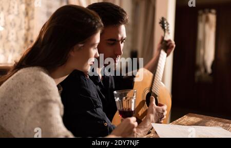 Giovane donna con vino guardando ragazzo leggendo foglio di carta e. suonare la chitarra mentre si crea la canzone in serata in un appartamento d'epoca Foto Stock