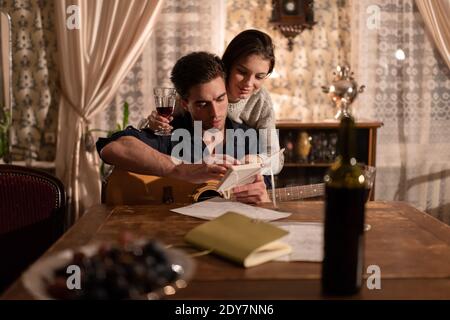 Giovane con chitarra e donna con bicchiere di vino controllo delle note nel blocco note durante la creazione di un brano vicino alla tabella in antico appartamento Foto Stock