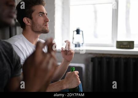 Maschio ansioso con una bottiglia di birra che attraversa le dita e sostiene Squadra preferita con un amico afro-americano mentre si guarda la partita insieme in ap moderno Foto Stock