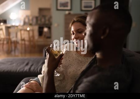 Felice giovane donna sorridente e aggraffante occhiali con ragazzo nero mentre gusterete il vino sul divano durante la data romantica in un ambiente accogliente camera Foto Stock