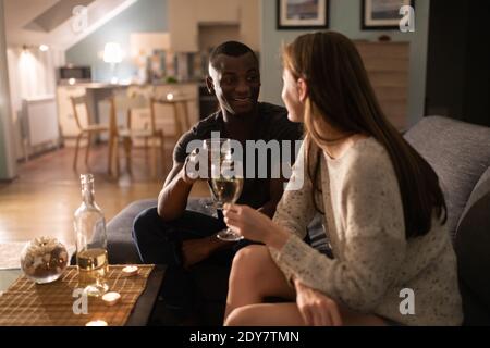 Ottimista afroamericano ragazzo sorridente e proponendo brindisi alla ragazza mentre si siede sul divano e si beve vino durante la data romantica a casa Foto Stock