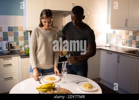 Afroamericano uomo limatura bicchieri con vino mentre giovane donna mettere piatto con spaghetti sul tavolo durante la data romantica a. casa Foto Stock