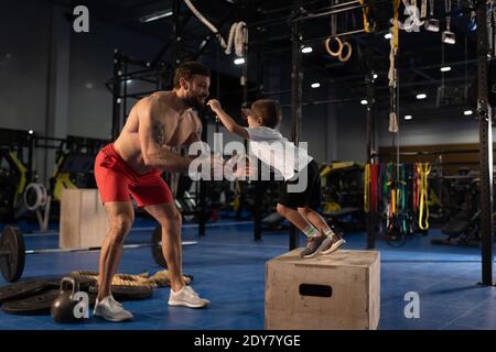 Il corpo pieno eccitava il portatore sorridente e catturante il ragazzo saltante mentre insegnano al figlio di essere forte durante l'allenamento fitness in palestra Foto Stock