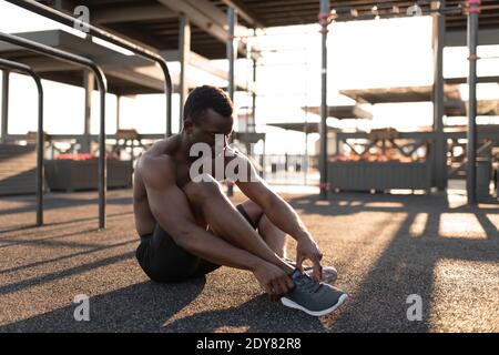 Atletico afroamericano uomo seduto e legando scarpe su sneaker per allenarsi alla luce del sole sul parco giochi Foto Stock