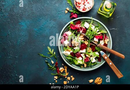 Insalata di barbabietole o barbabietole con rucola fresca, radicchio, formaggio morbido e noci sul piatto con forchetta, condimento e spezie su fondo blu del tavolo da cucina Foto Stock