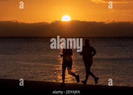 Southend on Sea, Essex, Regno Unito. 25 Dic 2020. Il giorno di Natale a Southend ha dato il via a freddo ma luminoso, con temperature appena al di sopra del gelo. I joggers sono fuori che corrono lungo le promenades all'alba. Due corridori femminili che si corrono lungo il mare mentre il sole sorge Foto Stock