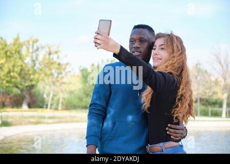 Uomo nero africano e rosso donna caucasica sorridente e prendendo un selfie con il loro smartphone in un parco. Giovane coppia multirazziale ritratto. Foto Stock