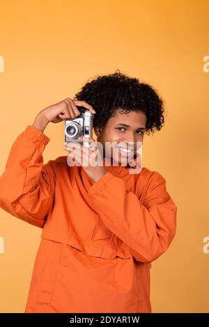 Un giovane moderno ed elegante, dalla pelle scura, sorride sinceramente, ponendosi sullo sfondo giallo di uno studio e scattando foto su una macchina fotografica vintage. C Foto Stock
