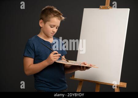 Mescolando le vernici ottenendo il colore desiderato che tiene una tavolozza con dipinge il ragazzo di 10 anni in t-shirt blu guarda fotocamera sullo sfondo di un cavalletto Foto Stock