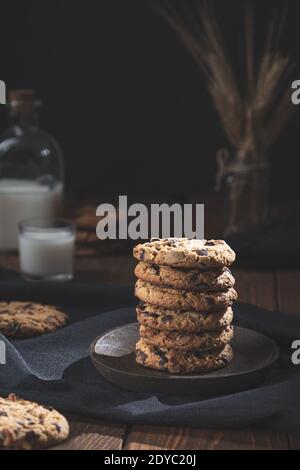 Biscotti al cioccolato accatastati, con un bicchiere e una bottiglia di latte su una base di legno, fondo scuro. Concetto di cibo dolce. Foto Stock