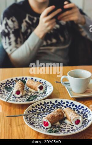 Cafe data mangiare italiano Dessert Cannoli. Uomo che tiene il telefono cellulare e pasta dolce fritta, farcita con ripieno cremoso di ricotta contenente ricotta Foto Stock