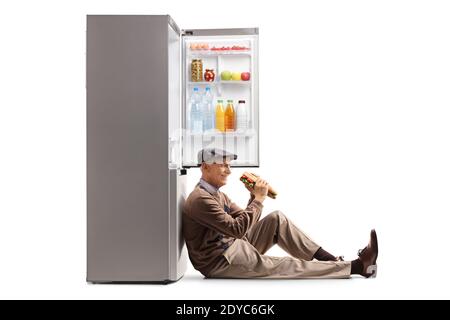 Uomo anziano che mangia un panino, seduto sul pavimento e appoggiato su un frigorifero aperto isolato su sfondo bianco Foto Stock
