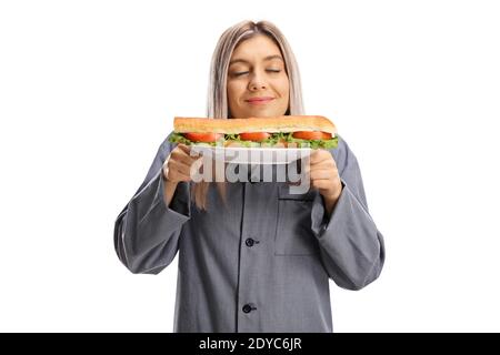 Donna affamata in pigiama che tiene un piatto con un panino in baguette e godendo con gli occhi chiusi isolati sopra sfondo bianco Foto Stock