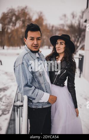 Uomo messicano ispanico in denim oversize giacca e donna in giacca biker in pelle di cappello che bacia il hugging ridendo matrimonio sposa walking.groom coppia d'amore Foto Stock