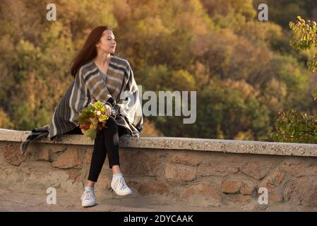 Bella giovane donna seduta sulla strada sullo sfondo della natura e pensando alla vita. Bouquet di foglie d'autunno in mano Foto Stock