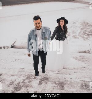 Uomo messicano ispanico in denim oversize giacca e donna in giacca biker in pelle di cappello che bacia il hugging ridendo matrimonio sposa walking.groom coppia d'amore Foto Stock