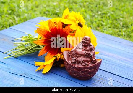 Statuina di buddha in ceramica su tavole di legno blu invecchiate nel parco estivo con fiori di rudbeckia. Foto Stock