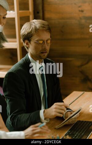 Giovane uomo con bicchieri freelance che lavora sul computer in ufficio. Uomo caucasico che mostra note ad un supervisore asiatico maturo. Operaio di collare bianco Foto Stock