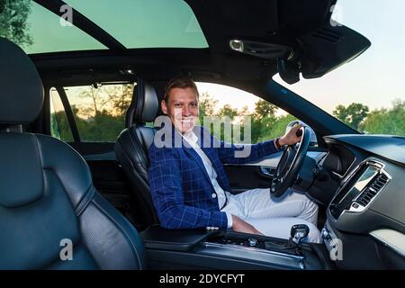 Un ragazzo in una giacca di plaid e pantaloni bianchi sorridente si siede al volante di un'auto costosa Foto Stock