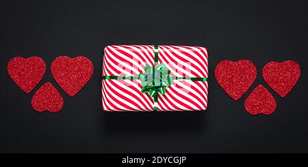 San Valentino, concetto d'amore. Romantica card panoramica scura con confezione regalo a righe bianche con arco verde. Regalo festivo e cuori rossi su una schiena nera Foto Stock