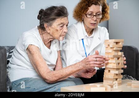 Jenga gioco. Il tema è la demenza, l'invecchiamento e i giochi per gli anziani. Caucasica anziana costruisce torre di blocchi di legno con l'aiuto di un medico come parte Foto Stock