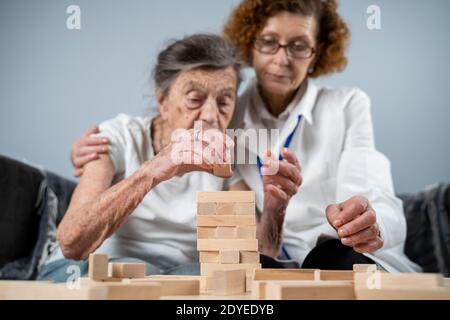 Jenga gioco. Il tema è la demenza, l'invecchiamento e i giochi per gli anziani. Caucasica anziana costruisce torre di blocchi di legno con l'aiuto di un medico come parte Foto Stock