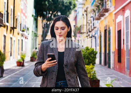 Donna messicana che tiene un telefono e che sta testando mentre è Camminando nella strada di una città dell'America Latina Foto Stock