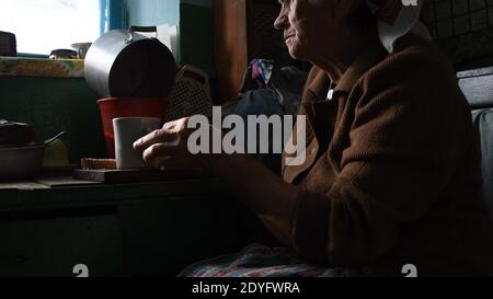 Donna anziana rugosa seduta dalla finestra della cucina e sofferente solitudine all'età avanzata. Ritratto di una donna rurale anziana nel contorno del sole. Concetto di p Foto Stock