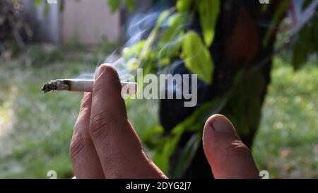 Closeup di fumo grigio volante da sigaretta che tiene in dita sporche di mano maschile su sfondo offuscato esterno. Cenere grigia all'estremità della sigaretta. Unhealt Foto Stock