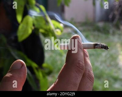 Closeup di fumo grigio volante da sigaretta che tiene in dita sporche di mano maschile su sfondo offuscato esterno. Cenere grigia all'estremità della sigaretta. Unhealt Foto Stock