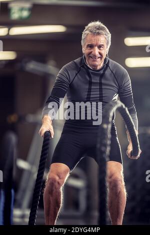 L'uomo anziano con le cuffie esercizi con le corde in un centro di allenamento fitness. Foto Stock