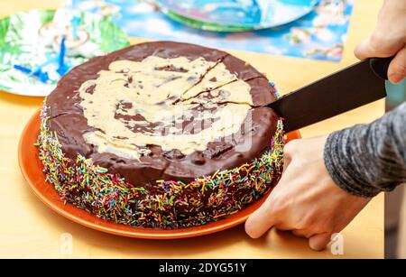 Persona che taglia, dividendo una torta di cioccolato fatta in casa decorata con molti spruzzi utilizzando un coltello, mano closeup. Pasticceria di cacao fatta in casa, torta di compleanno Foto Stock