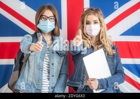 Due studentesse adolescenti che indossano maschere protettive mostrano i pollici in piedi davanti alla bandiera britannica. Foto Stock