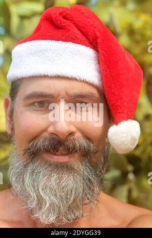 Un uomo bearded in un cappello di Santa guarda con sorriso alla macchina fotografica. Spiaggia tropicale, un thicket di cactus. Vista ravvicinata. Foto Stock