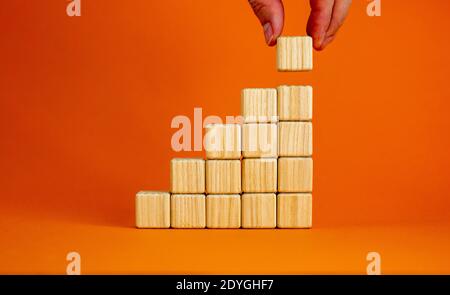 Simbolo della fondazione di successo della costruzione. Gli uomini mettono a mano un cubo di legno sulla piramide da blocchi di legno. Splendido sfondo arancione, spazio per la copia. Azienda A. Foto Stock