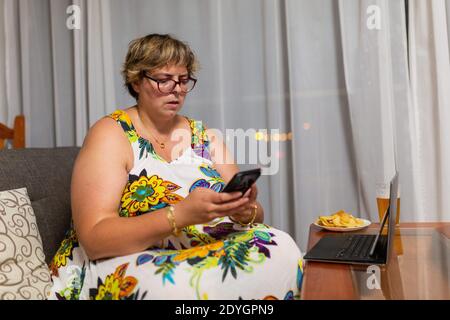 donna sovrappeso in un abito floreale seduto sul divano con un telefono cellulare in mano, laptop sul tavolo con un piatto di patatine e un bicchiere di birra Foto Stock