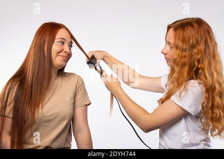 Le amiche stilizzano i capelli con una piastra per capelli. Concetto di cura dei capelli su sfondo bianco. Foto Stock