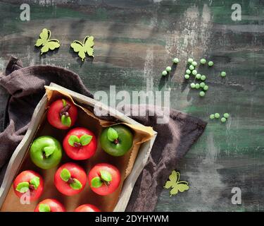 Mele spugne marzapane, verdi e rosse. Dessert creativo gustoso su vassoio di legno rustico con asciugamano di cotone tessile. Cibo creativo primaverile su texture scura Foto Stock