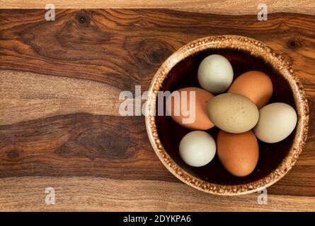 uno sfondo alimentare caratterizzato da una ciotola in ceramica di uova colorate su un tagliere di legno. Il lato sinistro è aperto per il testo. Foto Stock