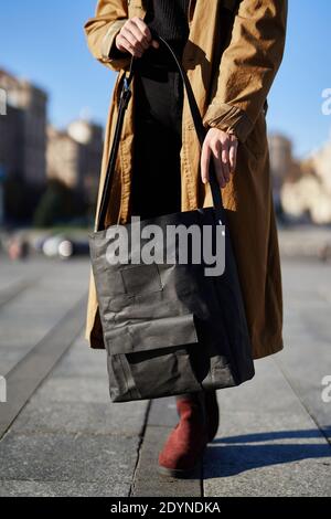 Nero eco carta fatta borsa nelle mani di una ragazza che indossa trincea arancione con cielo blu e grande città sfondo. Concetto di sacchetti di carta eco moda. Foto Stock