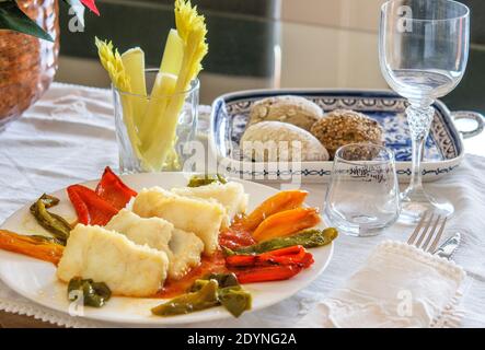 Grande piatto bianco con fette di merluzzo fritto e peperoni dolci, un piatto con vari panini e un bicchiere di verde sul tavolo decorato in festa. Foto Stock